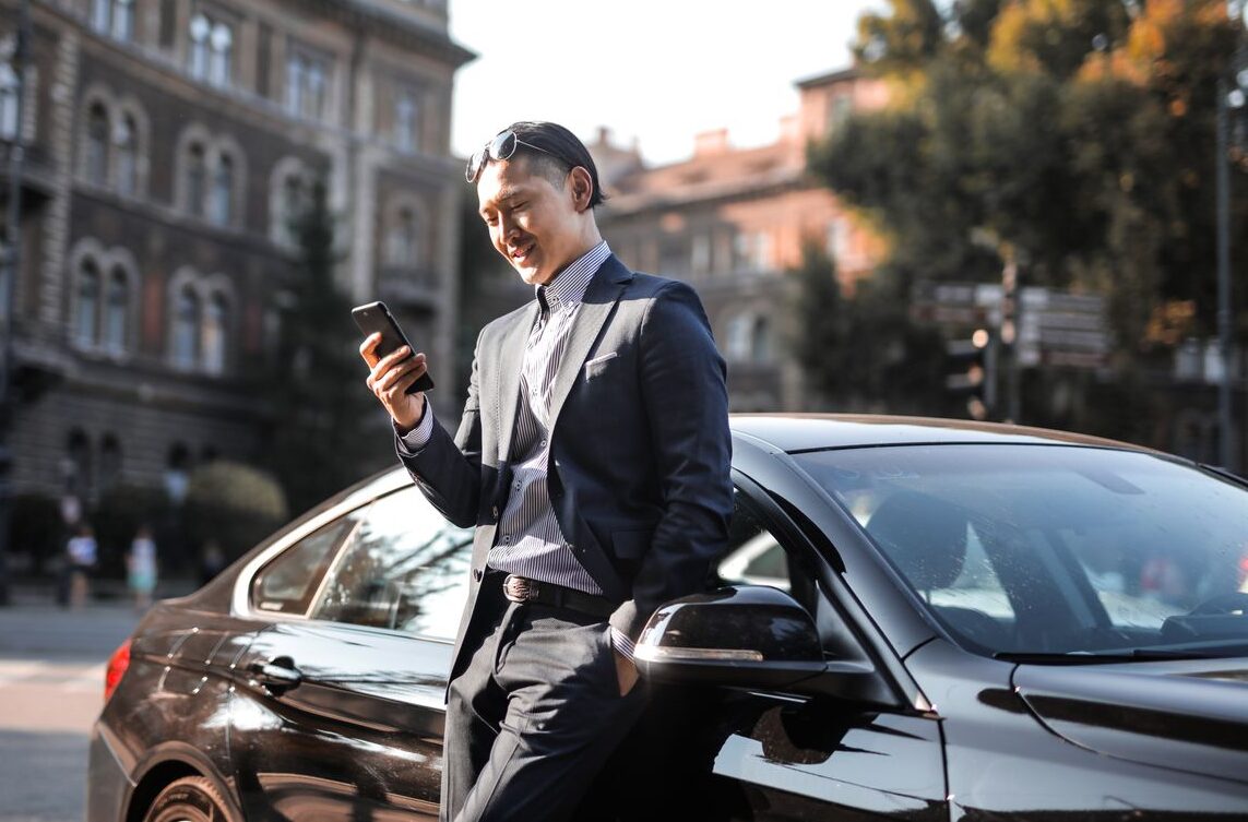 Smiling businessman in a black suit texting on his phone while leaning against a parked car in the city.