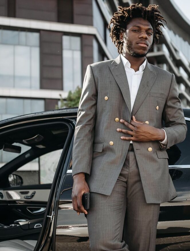 A confident businessman in formal attire stands by an elegant car in an urban setting.